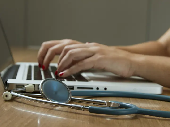Hands typing on a laptop beside a stethoscope on a desk