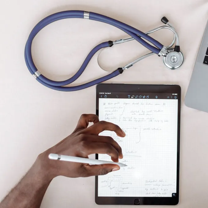 Overhead view of a desk with a stethoscope and a tablet where a hand takes handwritten notes with a stylus