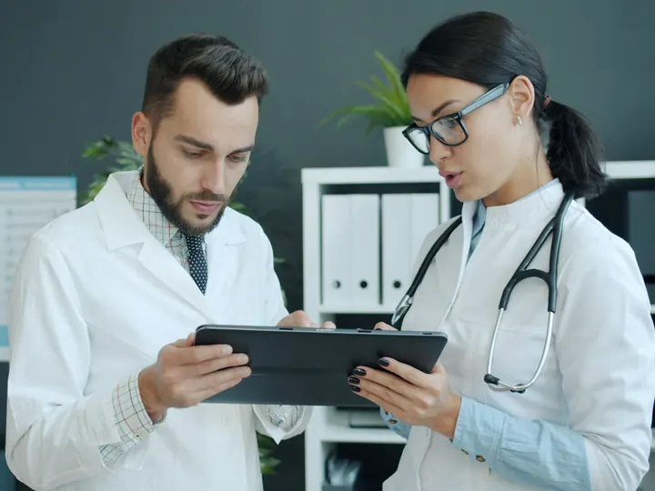 Two doctors in white coats reviewing information together on a tablet in a clinic office